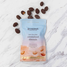 Load image into Gallery viewer, Pouch of dark chocolate caramelized almonds on white marble with chocolate-covered almonds scattered around, one split open.