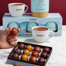 Load image into Gallery viewer, Cozy Season Gift Set: a hand holds a bonbon over a box of fall caramels, with hot chocolate mugs and mix cylinders behind.