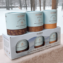 Load image into Gallery viewer, Classic Hot Chocolate Collection: Classic Dark, Mocha, and Classic Milk cylinders atop their boxed set by a snowy window
