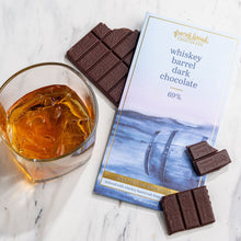 Load image into Gallery viewer, Whiskey Barrel Dark Chocolate 69% bar package beside a glass of whiskey over ice with dark chocolate pieces on marble.
