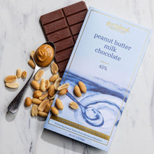 Load image into Gallery viewer, Peanut Butter Milk Chocolate 45% bar with wrapper, a spoonful of peanut butter, and scattered peanuts on marble.
