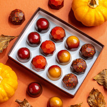 Load image into Gallery viewer, Open box of Fall Caramels Collection bonbons in red, maroon, and amber tones beside small pumpkins and dried autumn leaves.
