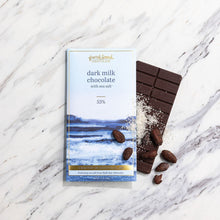Load image into Gallery viewer, Dark Milk Chocolate with Sea Salt 53% bar beside its blue wrapper, sprinkled with coarse salt and cacao beans on marble.
