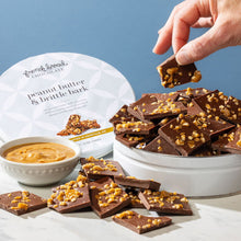 Load image into Gallery viewer, Hand lifting a square of peanut butter brittle bark from a full tin, with a bowl of peanut butter and pieces on marble.
