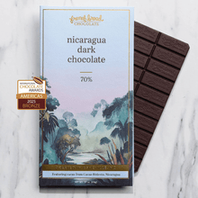 Load image into Gallery viewer, Nicaragua Dark Chocolate 70% bar next to its misty rainforest watercolor wrapper on a marble surface.
