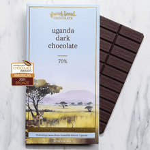 Load image into Gallery viewer, Uganda Dark Chocolate 70% bar beside its savanna watercolor wrapper with acacia trees on a marble surface.
